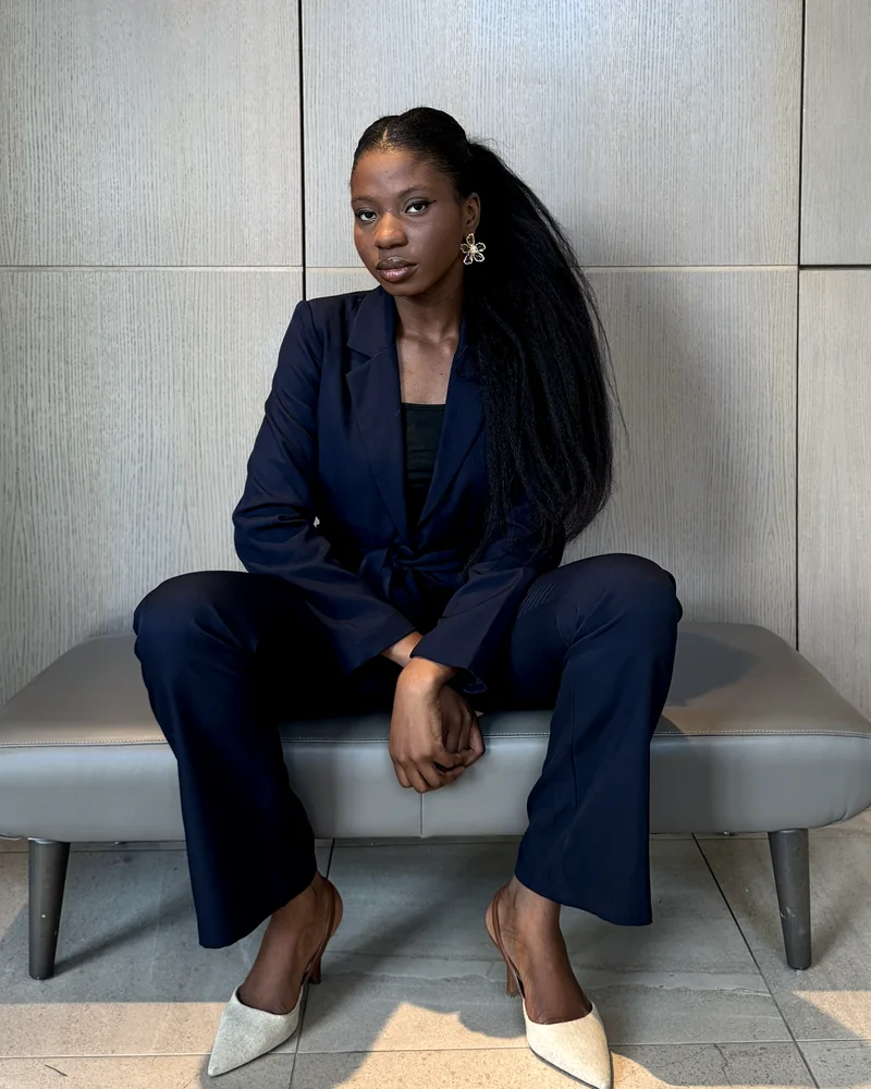 Maryann Eniola seated on a leather bench in a navy suit, looking at the camera